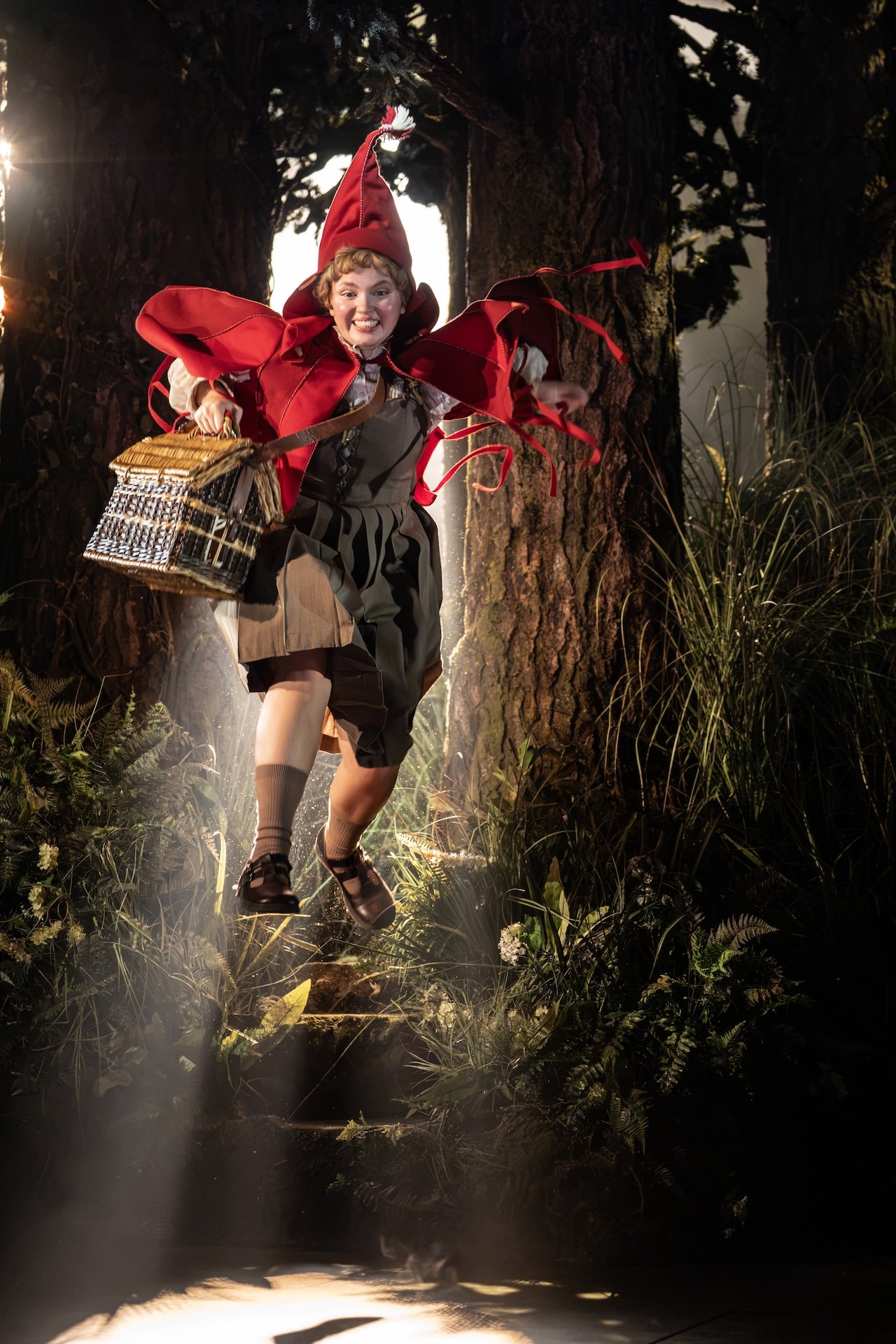Little Red Riding Hood leaps down forest steps mid-air, wearing a red cape and hood and carrying a wicker basket, in Into the Woods. Little Red Riding Hood leaps down forest steps mid-air, wearing a red cape and hood and carrying a wicker basket, in Into the Woods.