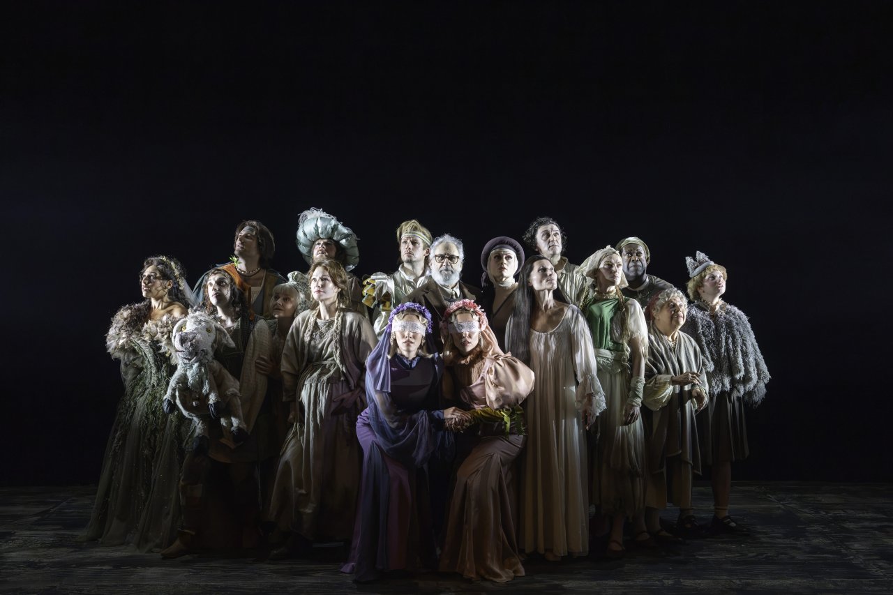 The company of Into the Woods stand together in muted fairytale costume against a black backdrop, two brides at the front with floral blindfolds, lit in a single spotlit tableau. The company of Into the Woods stand together in muted fairytale costume against a black backdrop, two brides at the front with floral blindfolds, lit in a single spotlit tableau.
