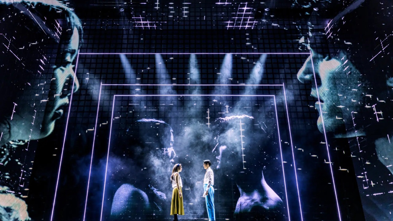 Two people standing on a stage with their images projected on a screen. Maybe Happy Ending. Broadway. Photo by Matthew Murphy and Evan Zimmerman.