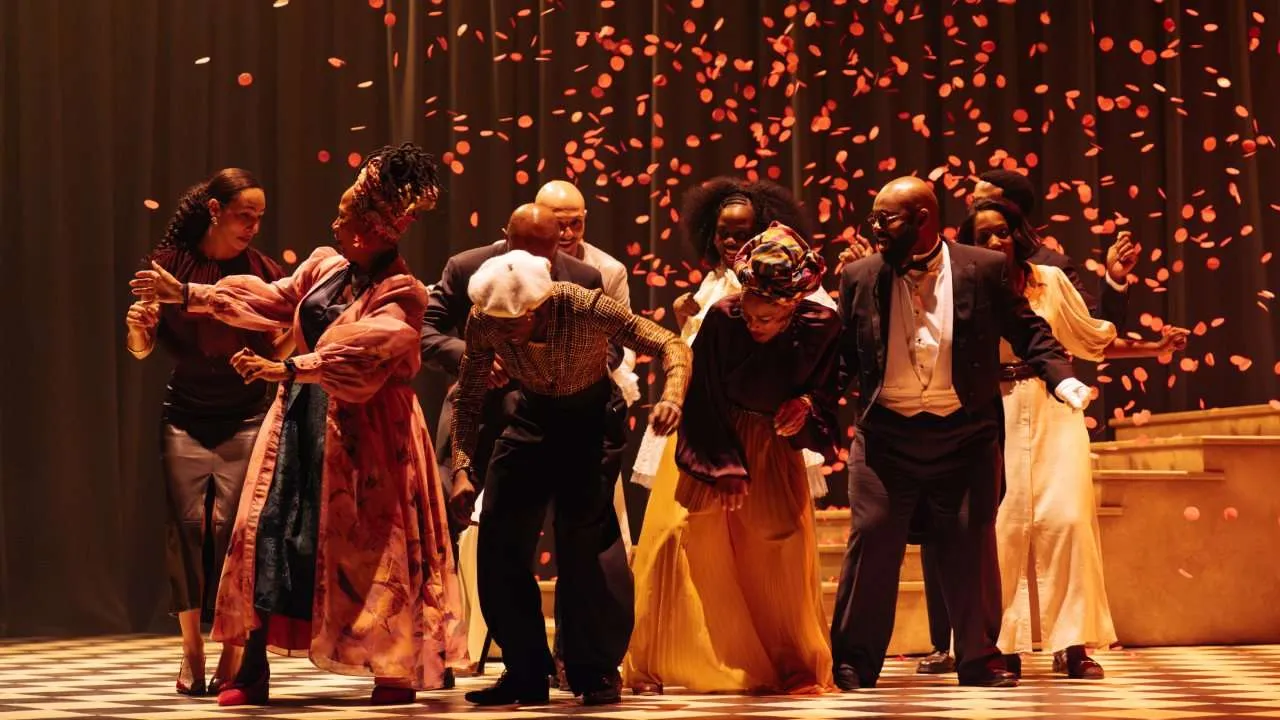 Diverse group of dancers performing on a checkered stage as orange petals fall around them, in warm lighting.