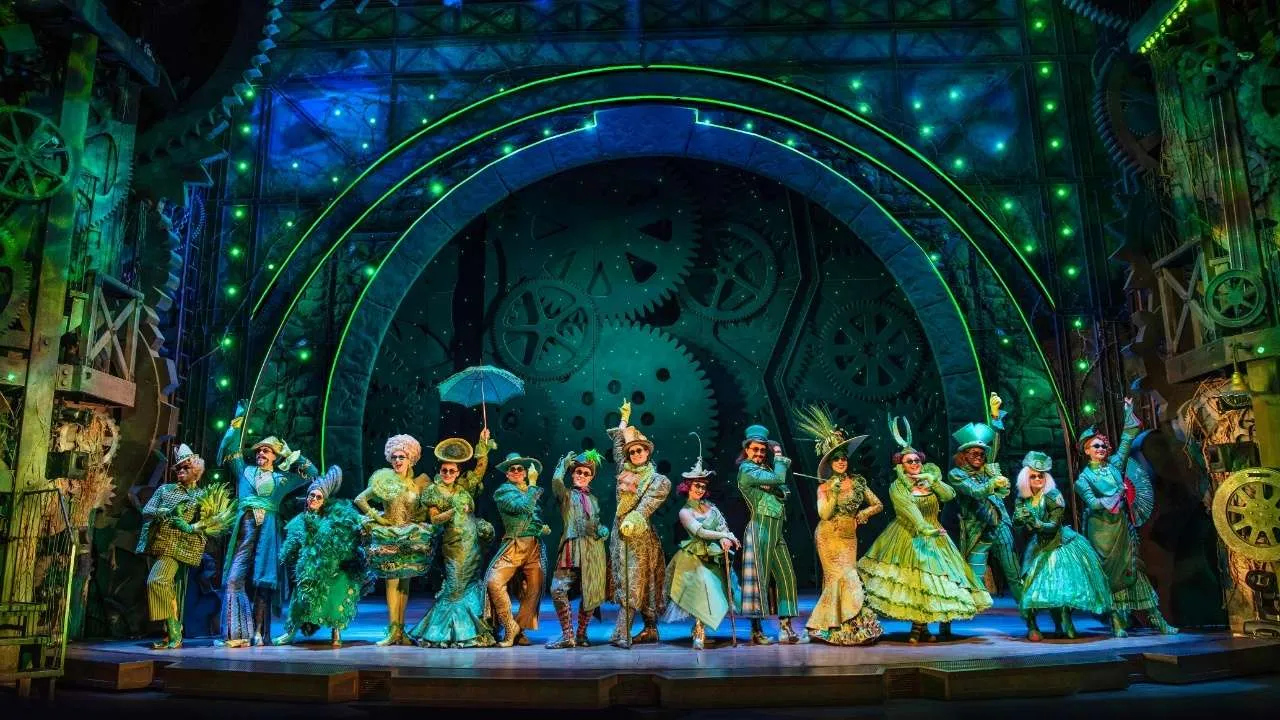 Line of performers in steampunk and vintage costumes posing on a brightly lit theater stage with a gear-themed backdrop and neon green rings.