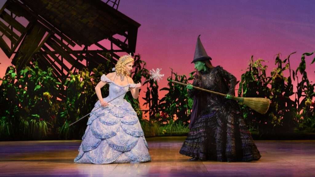 Two women face off on a stage. One, Glinda, wears a sparkly blue dress and a tiara, and brandishes a septre. The other, Elphaba, has green skin and wears all black. She brandishes a broom.