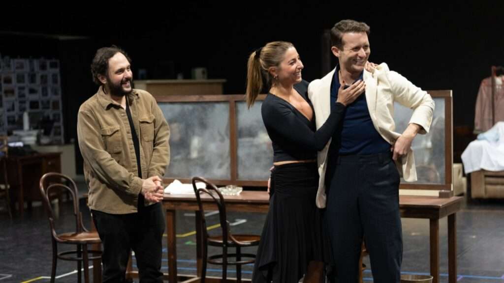 Three performers on a stage, smiling; two actors embrace while a man in a tan jacket watches warmly nearby.