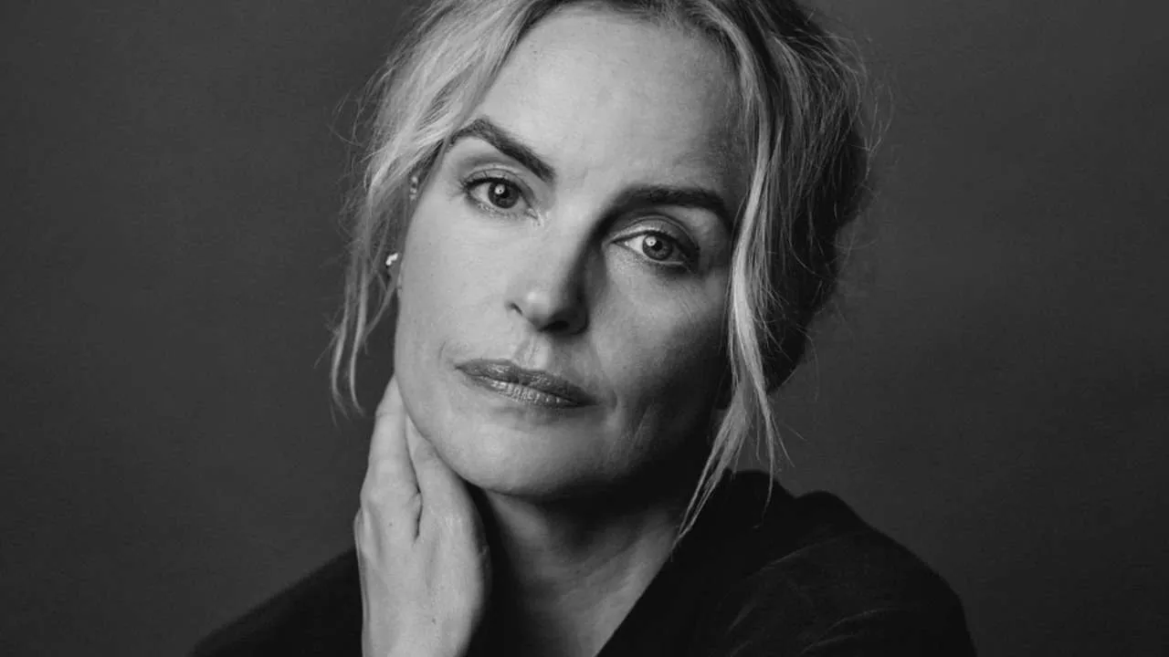 Black-and-white portrait of a woman with light hair, hand touching her neck, looking at the camera.