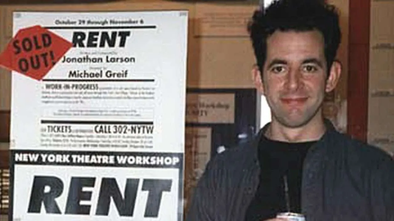 Man standing beside a large RENT theatre poster with a red SOLD OUT sticker