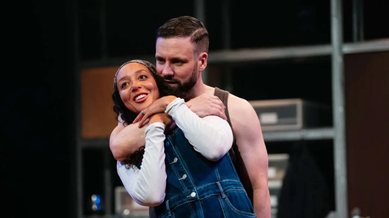 Two actors on a stage share a warm hug: a smiling woman in denim overalls and a headband is embraced from behind by a bearded man in a sleeveless shirt, backdrop of rehearsal space visible behind them.