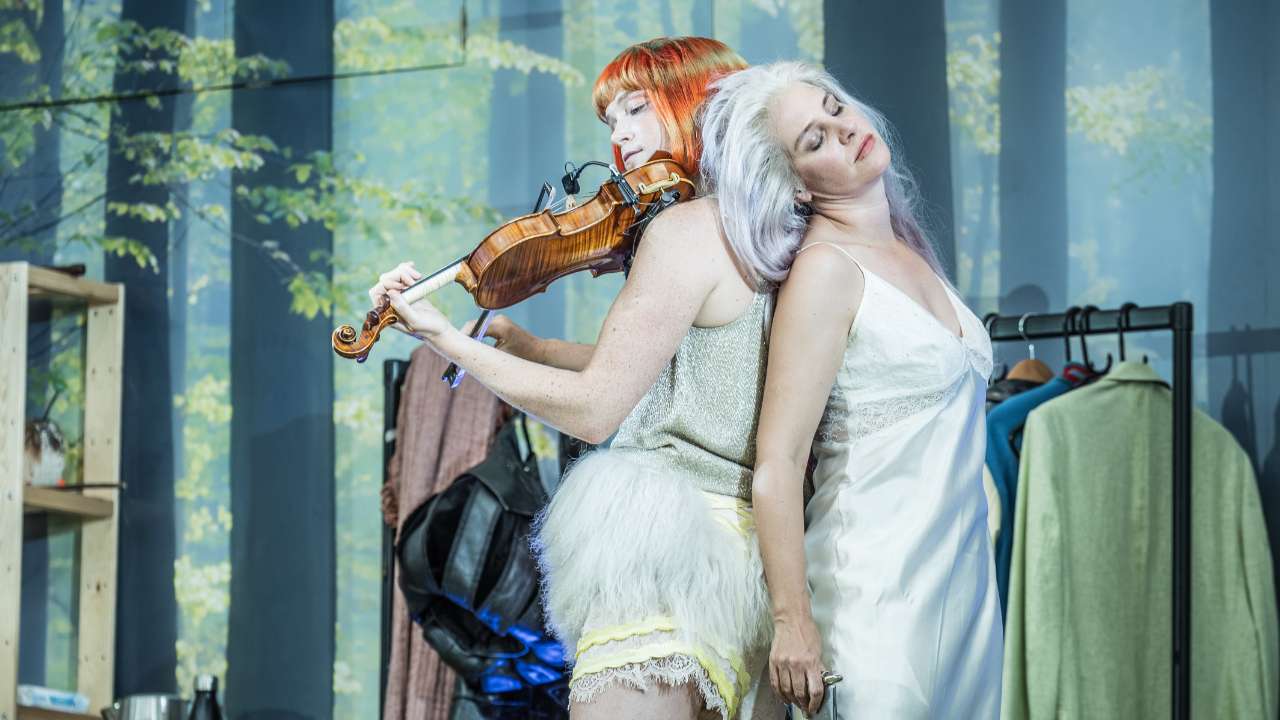 Two women back to back backstage: a violinist with orange hair in a silver top and fluffy white skirt, and a silver‑haired woman in a white satin dress resting her head on her shoulder.