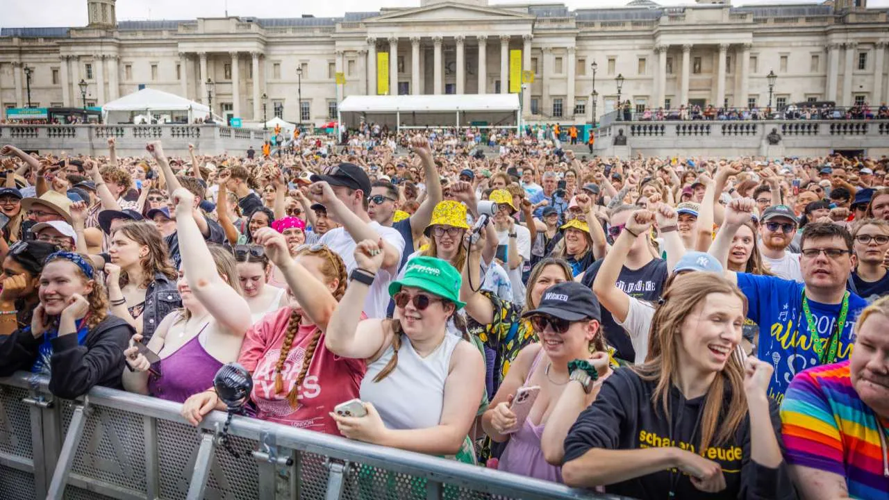 Large diverse crowd at an outdoor festival, with arms raised and smiling, in front of a neoclassical building and stage tents.