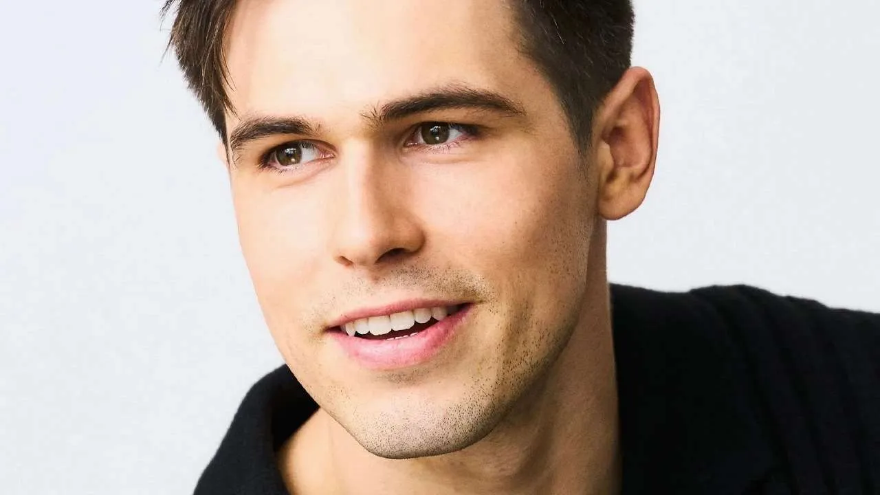 Close-up portrait of a smiling man with short brown hair and light stubble, wearing a black shirt against a pale background.