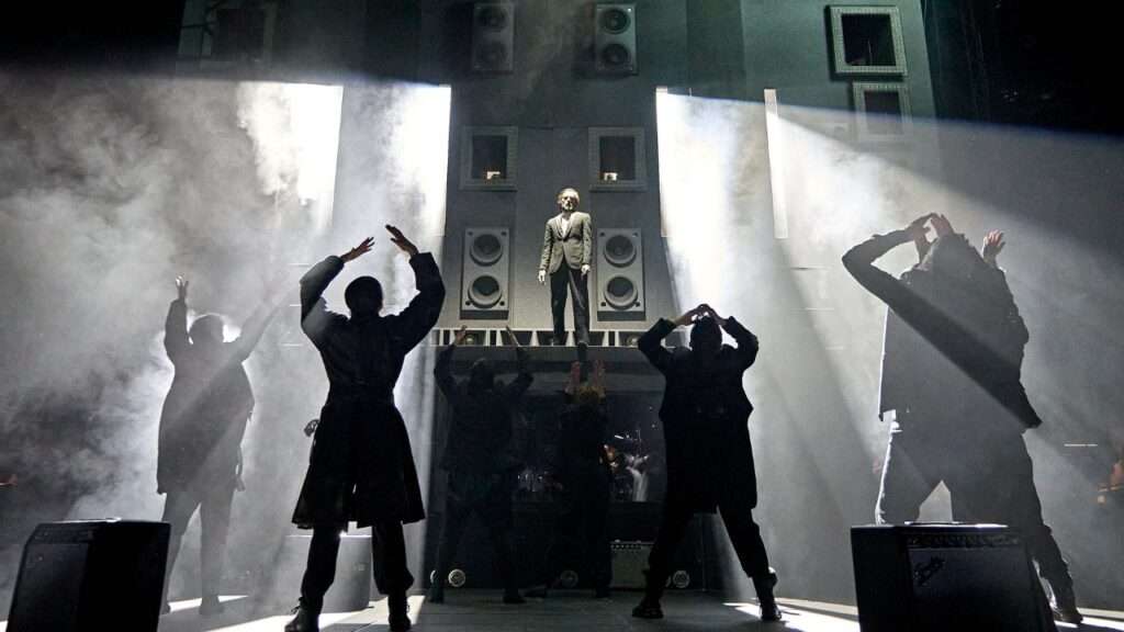 Male performer in a suit stands on a raised platform amid speakers and fog, performing on a dark concert stage with silhouettes of fans raising arms in front.