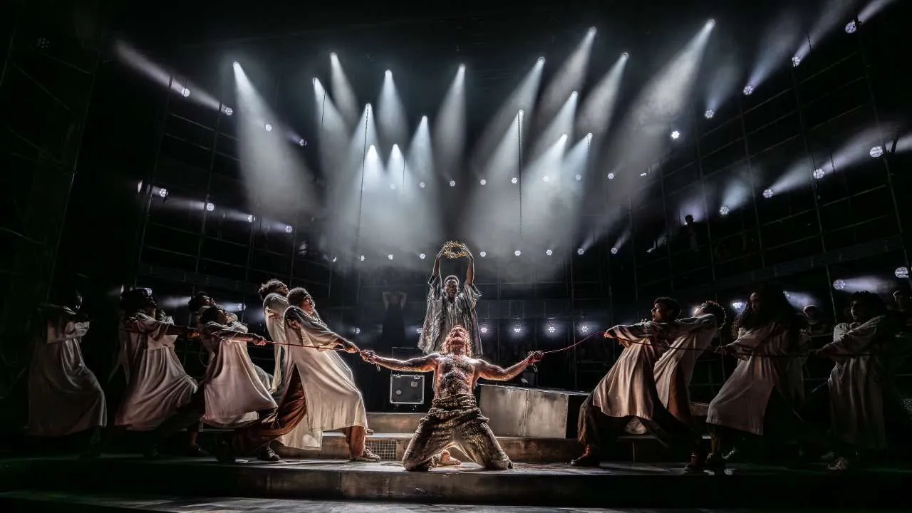 Theater stage scene: a central kneeling dancer with arms outstretched, surrounded by dancers in light robes pulling ropes under dramatic spotlights.