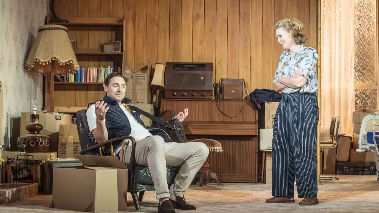 Man relaxing in a retro wood-paneled living room, gesturing as a woman stands nearby in striped pants amid boxes and vintage furniture.
