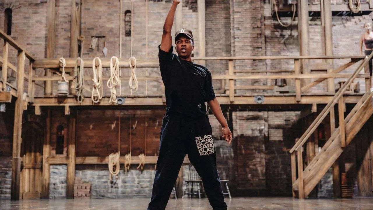 Man in a black outfit dances with one arm raised inside a wooden, rope-filled rehearsal space.