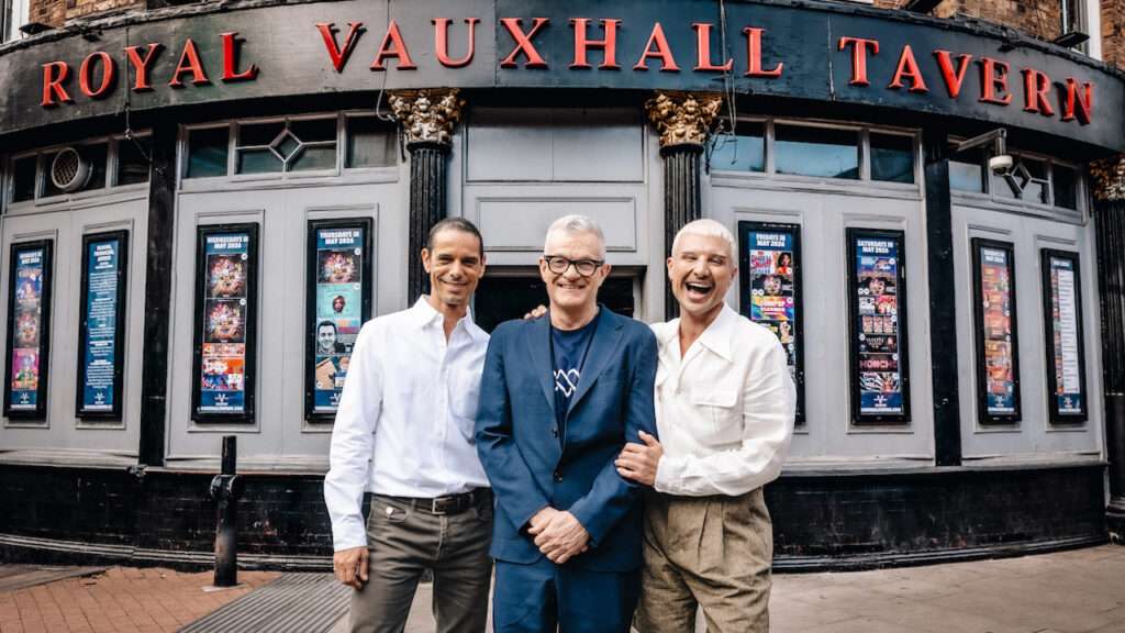 Three men pose and smile in front of the Royal Vauxhall Tavern outside a pub with red letters on the sign.
