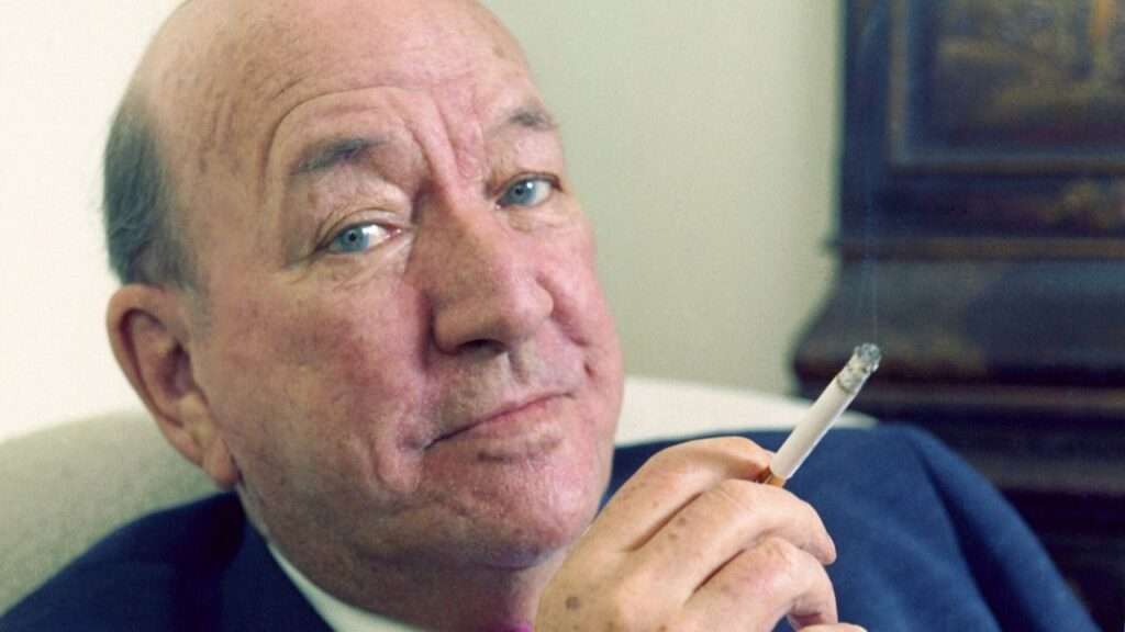 Close-up of a smiling older man with blue eyes holding a lit cigarette outside his lips, looking at the camera.