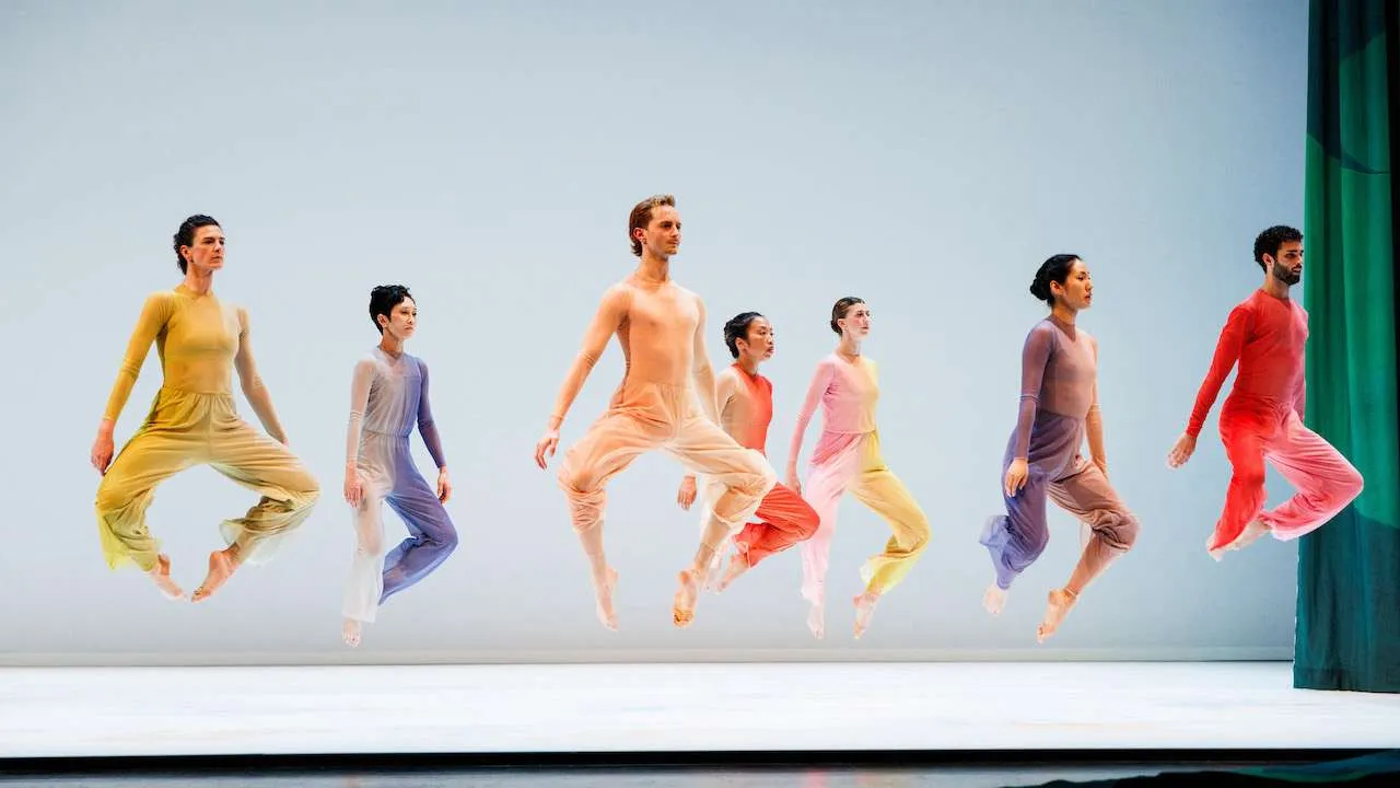 Group of dancers in colorful outfits leaping in midair on a minimalist stage with a pale blue background, synchronized across the line of performers.
