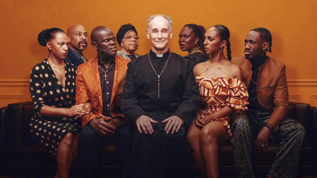 Priest in black robes sits center, flanked by seven well-dressed adults on a sofa against an orange wall.
