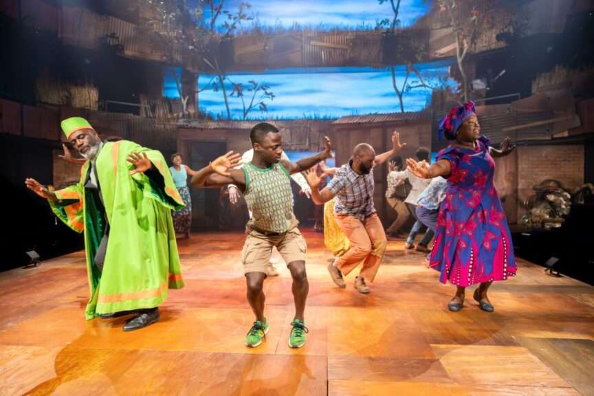 Diverse group of dancers performing energetically on a wooden stage in colorful traditional outfits, a bright green robe and a purple dress with patterns, mid-dance with arms extended.