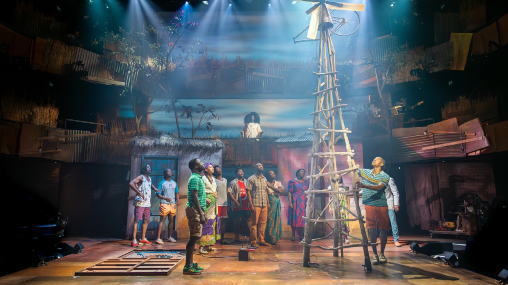 Theater stage with a tall wooden ladder-like scaffold at center, a group of diverse performers gathered around and looking up toward a performer on a balcony above the set.