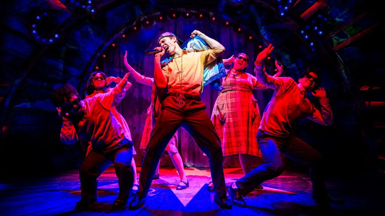 Performer center stage singing into a microphone while dancers pose dynamically under blue and red stage lighting on a wooden set.
