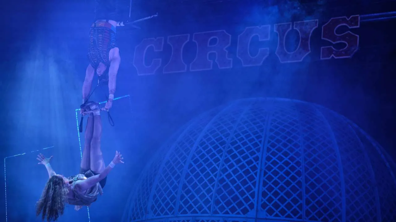 Aerial circus performers under blue lighting: one upside down on straps above another dancer holding below, with a lattice dome backdrop and the word CIRCUS visible.