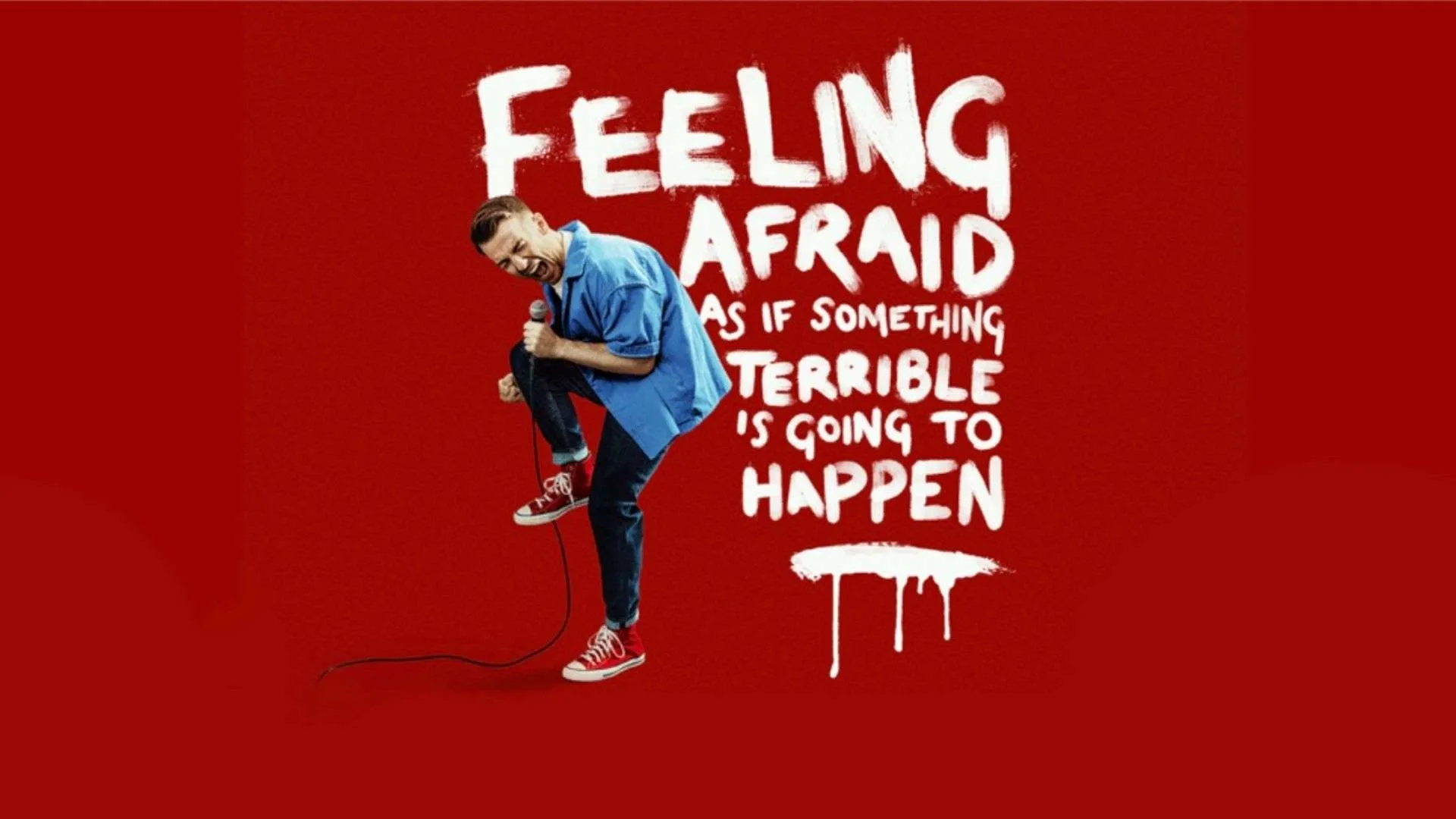 Man in a blue shirt and red sneakers sings into a microphone against a red background with white graffiti-style text nearby.