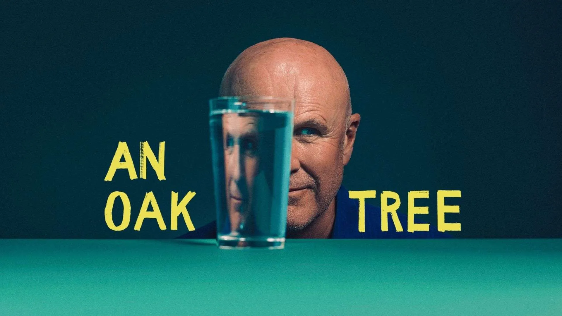 Man's face partly obscured by a tall glass of blue water on a teal table, with bold yellow text reading 'AN OAK TREE' in the background