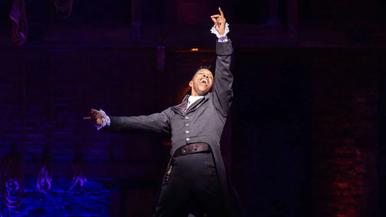 Male performer on stage joyfully raising one arm, other extended, wearing a 19th-century coat and ruffled cuffs amid blue and purple lighting.