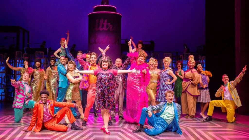 Large cast of performers in vibrant 1970s-style costumes posing on a theater stage.