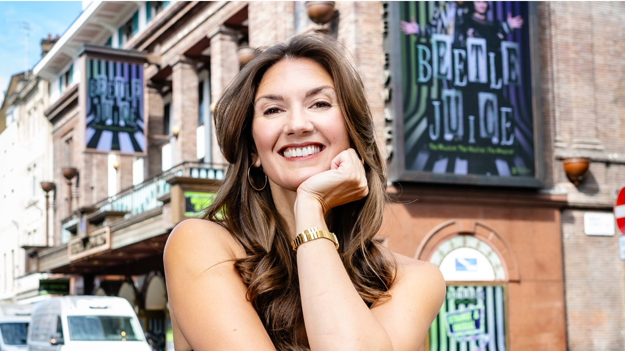 Smiling woman with hand under her chin posing on a city street with brick buildings and theater posters in the background, sunny day.