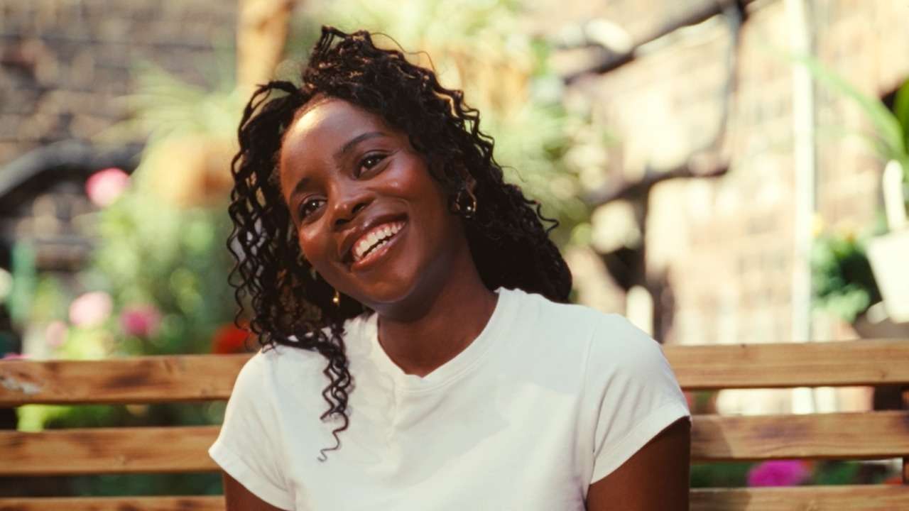 Portrait of a smiling Black woman sitting on a wooden bench outdoors, wearing a white t-shirt.