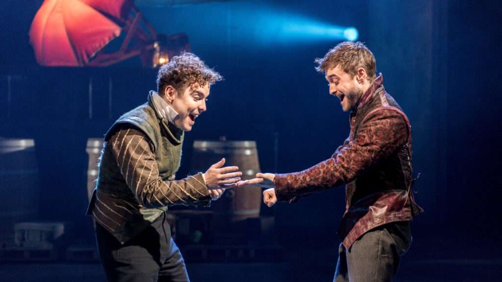 Two actors on a theater stage laugh as they reach toward each other, preparing to shake hands.