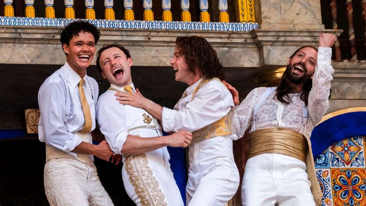 Four men in white costumes with gold sashes laugh and pose on a decorated stage, celebrating together.