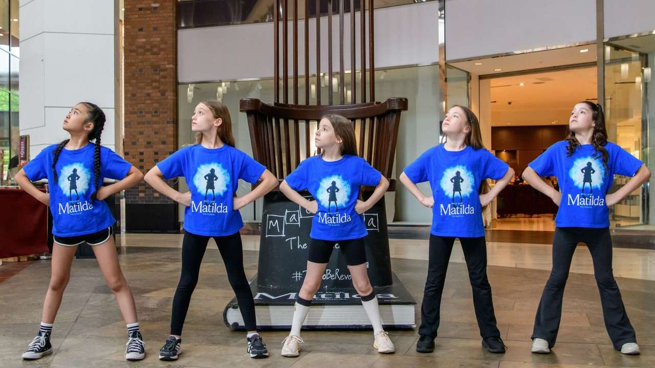 Five girls in blue Matilda T-shirts stand in a mall atrium with hands on hips, lined up and facing different directions.