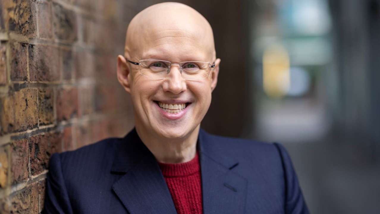 Smiling bald man in glasses wearing a navy blazer and red sweater, leaning against a brick wall.