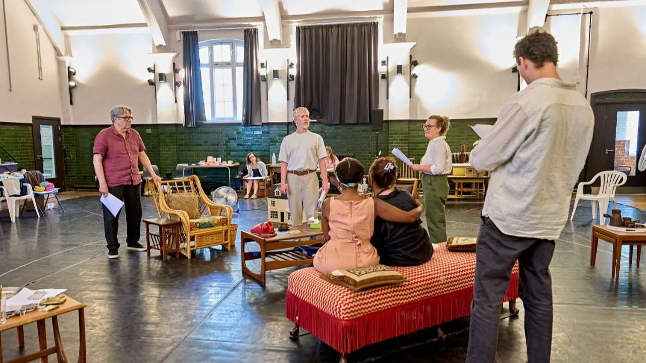 Actors rehearse a scene in a large hall filled with vintage furniture; two children sit on a red checkered sofa while adults hold scripts nearby.