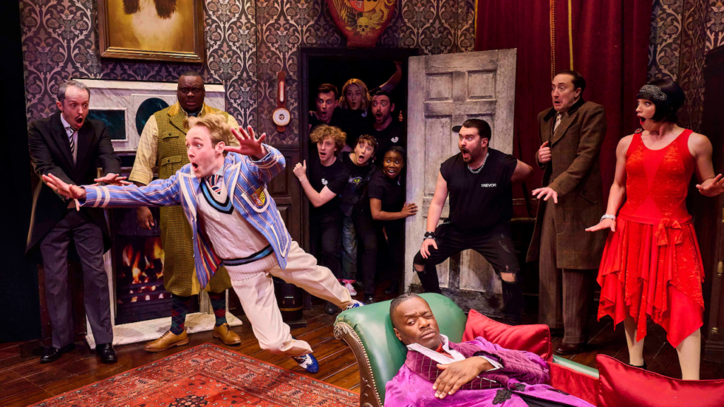 Stage moment: a man in a blue striped blazer lunges forward as fellow actors react with shock in a decorated set.