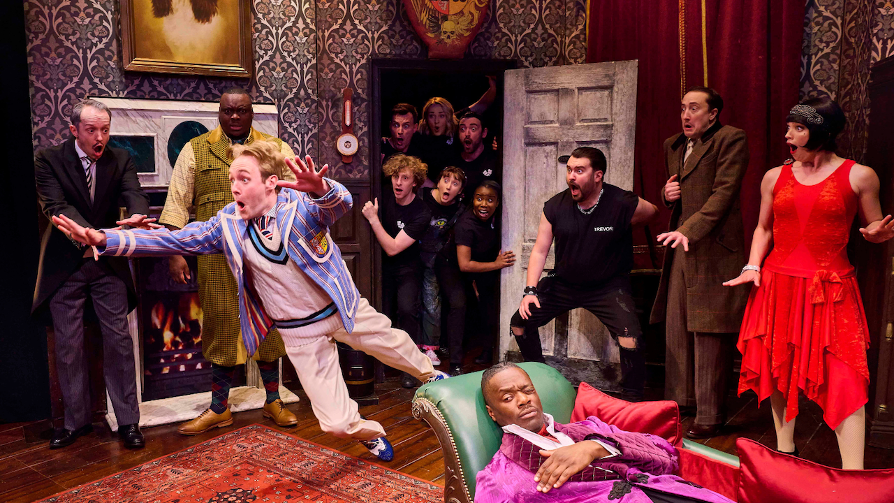 Stage moment: a man in a blue striped blazer lunges forward as fellow actors react with shock in a decorated set.