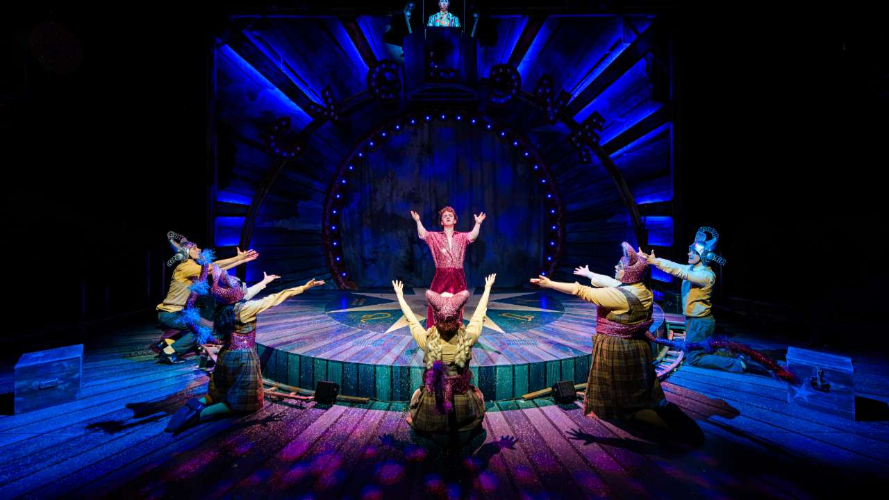 Circus-style stage with a central performer in pink, arms raised, surrounded by costumed kneeling performers under blue lights.