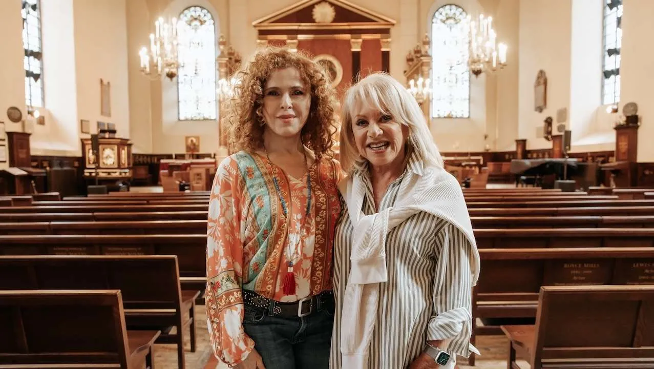 Two women stand together in a church nave, smiling at the camera among wooden pews and chandeliers with stained glass windows in the background.