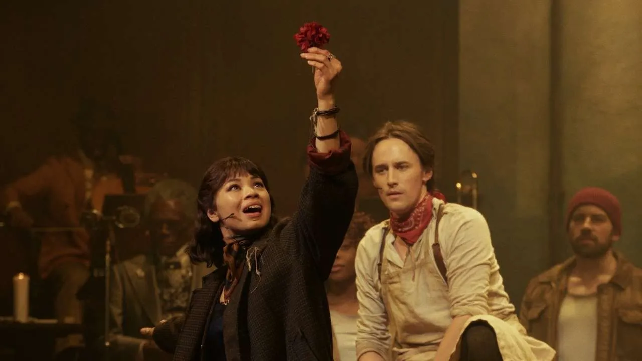 Woman on stage raises a red flower toward the audience as others watch in a dim, warm-lit theater.
