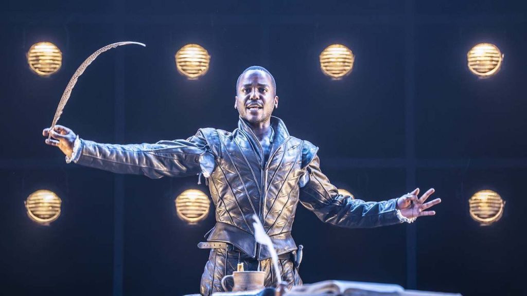 Male performer in a metallic jacket twirling a whip on stage under warm spotlights.