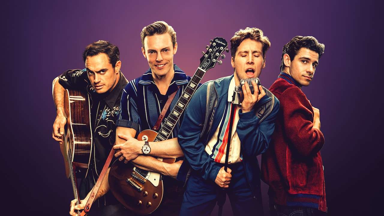 Four male musicians posing with guitars in a lineup against a purple background, one singing into a harmonica and another wearing a striped shirt and blue jacket with a watch visible on his wrist.