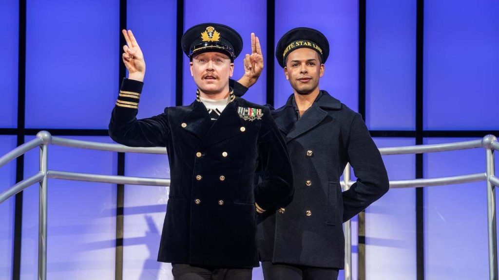 Two men in naval uniforms pose on a blue-lit stage; the left man raises a V for victory with both hands while the other stands beside him.