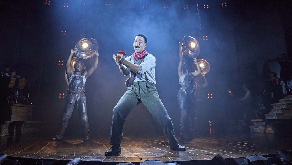 Male singer center stage, passionately performing with red rose in hand, flanked by two backup dancers holding circular stage lights in a smoky, blue-lit theater scene.