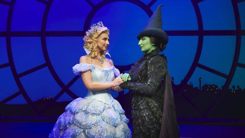 Two costumed performers share a handshake on stage: a princess in a light blue gown with a tiara and blonde curls, and a green-skinned witch in a tall hat and black dress, set against a blue circular window backdrop.