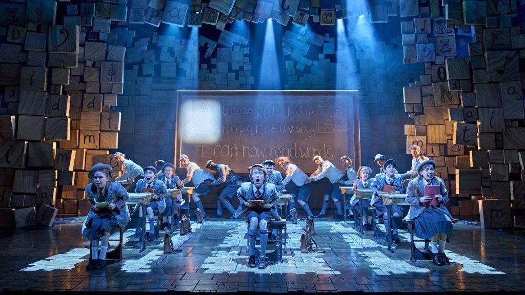 Children in school uniforms perform a classroom scene on stage, with a chalkboard reading 'I can now read words'.