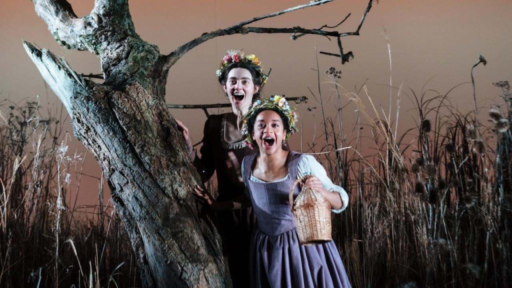 Two women in vintage dresses with flower crowns, hiding behind a large tree in a dry grassy field, one holding a wicker basket.