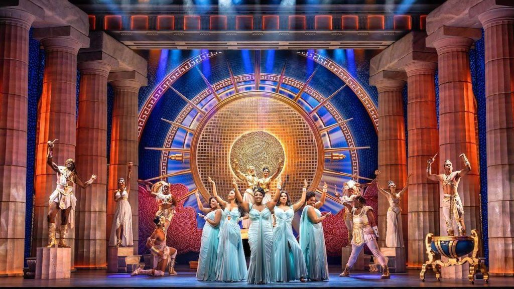 Group of dancers in light blue gowns perform a choreographed number on a grand classical-stage set with gold sunburst backdrop.