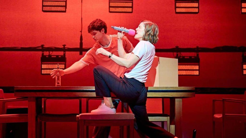 Woman singing into a pink microphone at a red-lit stage, with a man leaning on a table holding a bottle nearby.